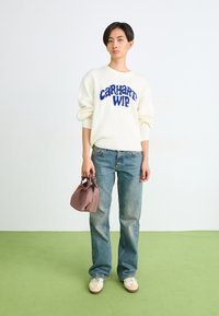 Kreemist sviitrimütsist sinise "Carhartt WIP" logoga, kantud helesiniste teksadega, hoides väikest mauve värvi käekotti ning beežide tossudega rohelisel põrandal.