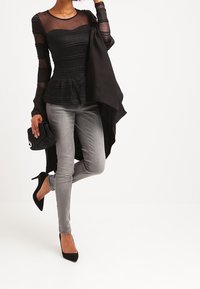 Haut noir semi-transparent à manches longues avec texture froncée, associé à un jean skinny gris et des escarpins noirs, tenant un clutch noir.