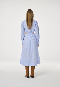 Vrouw met lang haar in een lichtblauwe midi-jurk met zijcut-outs en bruine suède kniehoge laarzen, staande op een witte achtergrond.