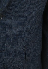Marineblauw stof met een visgraatpatroon en een gestructureerde afwerking, voorzien van een enkele zwarte knop en een zijzakdetail.