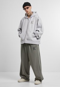 Hanorac gri cu fermoar și logo, asortat cu pantaloni de trening largi, de culoare verde măsliniu, cu logo. Purtați cu adidași de culoare crem.