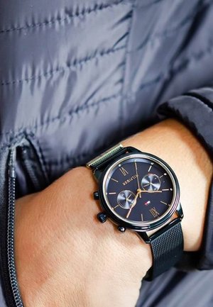 Montre chronographe noire ronde avec un bracelet en maille métallique, des accents dorés et un cadran sombre affichant trois sous-cadrans et des chiffres romains.