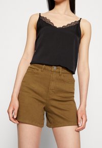 Camisole noire avec bordure en dentelle associée à un short marron, doté d'une taille haute, de cinq poches et de passants pour ceinture. Les matériaux semblent doux et décontractés.