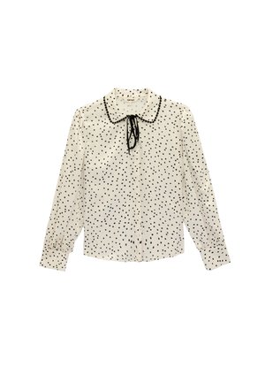 Valkoinen pitkähihainen pusero, jossa mustia epäsäännöllisiä pilkkuja, pyöreä kaulus, jonka reunoilla mustia helmiä, ja musta solmio kaula-aukossa.