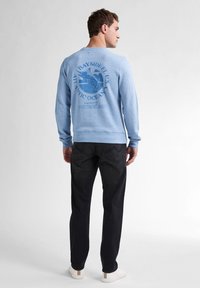 Lichtblauw sweatshirt met een cirkelvormige grafiek op de rug, met een vis en tekst, gecombineerd met donkere jeans en witte schoenen.