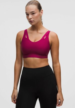Mujer con cabello rubio recogido en una cola de caballo, vistiendo un sujetador deportivo magenta y leggings negros de talle alto sobre un fondo claro y liso.