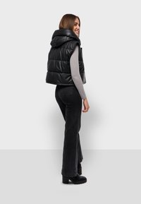 Gilet imbottito nero con cappuccio, realizzato in materiale lucido, presenta un design cropped e un motivo trapuntato, indossato sopra una maglietta grigia chiara e jeans scuri.