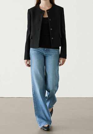 Femme portant une veste noire boutonnée sur un haut noir, un jean large bleu clair ample et des chaussures noires pointues, debout sur un sol neutre.