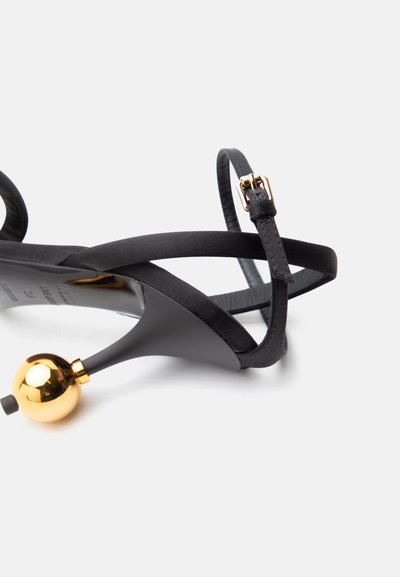 Μαύρο σανδάλι με ψηλό τακούνι και λεπτές λουρίδες, χρυσή αγκράφα και μοναδικό σφαιρικό χρυσό τακούνι σε λευκό φόντο.