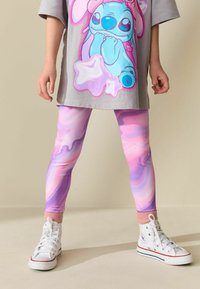 Szara, oversize koszulka z kolorowym nadrukiem Stitcha, połączona z różowo-fioletowymi legginsami marmurkowymi oraz białymi trampkami na wysokiej podeszwie z różowymi akcentami.