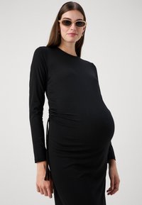Vestido preto de manga longa com franzido lateral, feito de um tecido suave. A modelo usa óculos de sol ovais castanhos; design minimalista.