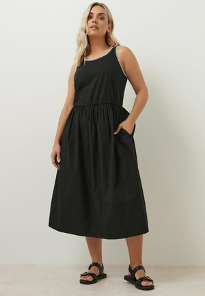 Schwarzes ärmelloses Kleid mit tailliertem Schnitt und gerafftem Rock. Es hat einen weißen Besatz am Ausschnitt und seitliche Taschen.