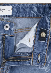 Elastični pojas s patent zatvaračem koji je djelomično otvoren, rupica za gumb s podesivim gumbom i oznakom "Regular" na unutarnjoj strani pojasa.