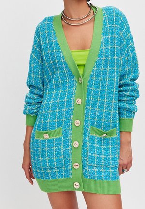 Woman wearing a blue and green buttoned knit cardigan with two front pockets over a lime green top and a silver necklace.