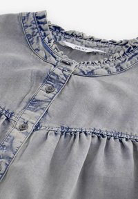 Blauwe denim top met een gerimpelde halslijn, knoopsluiting en geplooide taille-inleg. Heeft een vervaagde textuur en subtiele lichtblauwe patronen.