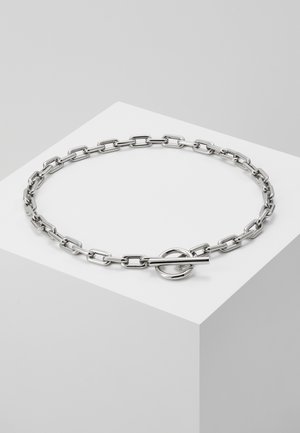 Zilverkleurige metalen schakelenketting met rechthoekige schakels en een ronde schakelsluiting op een wit voetstuk tegen een grijze achtergrond.