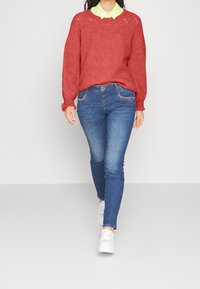 Rode gebreide trui met ronde hals, over een lichtgroen overhemd met kraag, gecombineerd met blauwe skinny jeans en witte sneakers.