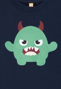 T-shirt in cotone blu con un mostro cartoon verde con una bocca larga, denti affilati e corna rosse, su uno sfondo blu navy scuro.