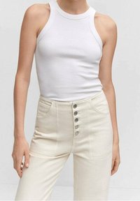 Persona que lleva una camiseta sin mangas blanca ajustada y pantalones crema de cintura alta con cinco botones de metal visibles en la parte delantera.