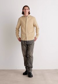 Camicia beige con bottoni e maniche lunghe, abbinata a pantaloni grigi e scarpe nere, in piedi su uno sfondo neutro.