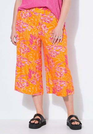 Mujer con pantalones anchos de color naranja brillante con estampados de hojas tropicales rosas, combinados con sandalias negras con hebilla y una camiseta rosa.
