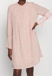 Długa sukienka z długim rękawem w jasnoróżowym materiale z rozproszonym ciemnoróżowym wzorem w panterkę, z plisami, okrągłym dekoltem i kieszeniami po bokach.