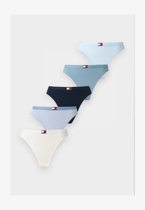 Frau mit langen dunklen Haaren, die einen weißen Tommy Hilfiger Bralette und blaue Unterwäsche trägt, steht vor einem schlichten hellen Hintergrund.