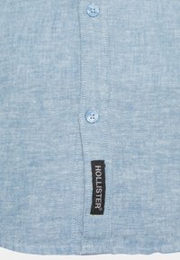 Chemise bleu clair à boutons, en tissu texturé, ornée de boutons bleus et d'une étiquette noire avec du texte blanc, située sur le côté gauche.
