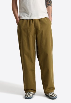 Person iført løstsiddende brune bukser, en hvid t-shirt og sort-hvide ternede slip-on sko med hænderne i lommerne.