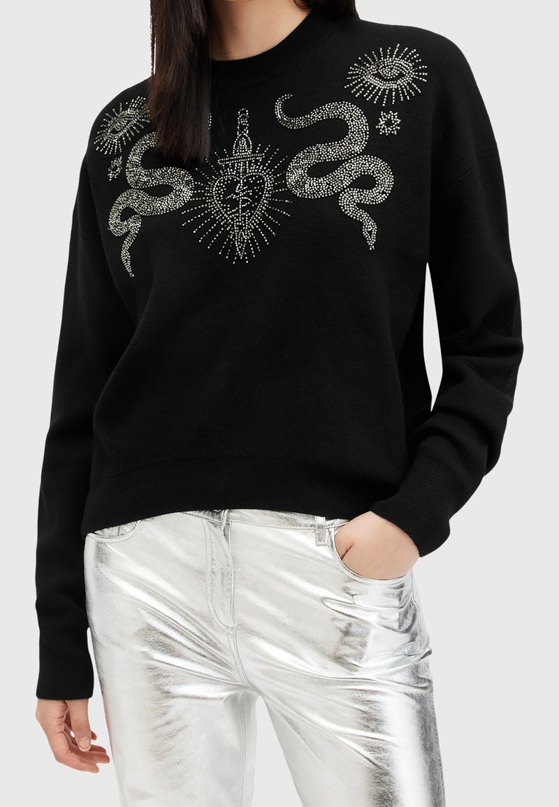 Personne portant un pull noir avec des motifs en strass représentant un serpent, un cœur et un œil, associé à un pantalon métallique argenté brillant.