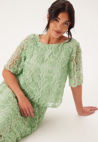 Top in pizzo floreale verde con maniche corte, caratterizzato da motivi di rose in rilievo e un sottile strato trasparente. Tessuto morbido e leggero, vestibilità rilassata.