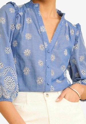 Niebieska bluzka z białym kwiatowym haftem oczkowym, falbankowym kołnierzem, rękawami o długości 3/4 z bufkami i guzikami pokrytymi tkaniną, noszona z białymi spodniami.