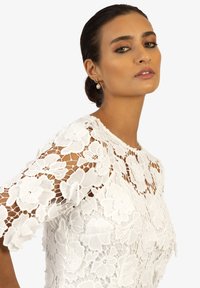 Weiße Spitzenbluse mit floralem Stickerei, kurzen Ärmeln, rundem Ausschnitt, strukturiertem Stoff, mit tailliertem Schnitt und transparenten Details.