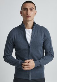 Mørkegrå cardigan med glidelås laget av mykt strikket materiale; har høy krage, ribbestrikkede mansjetter og hem. Modell som viser plagget.