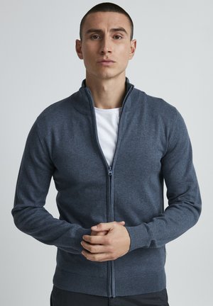 Mørkegrå cardigan med glidelås laget av mykt strikket materiale; har høy krage, ribbestrikkede mansjetter og hem. Modell som viser plagget.