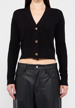 Mujer con un cárdigan negro corto de canalé con grandes botones y pantalones de cuero negros de talle alto, de pie frente a un fondo liso.
