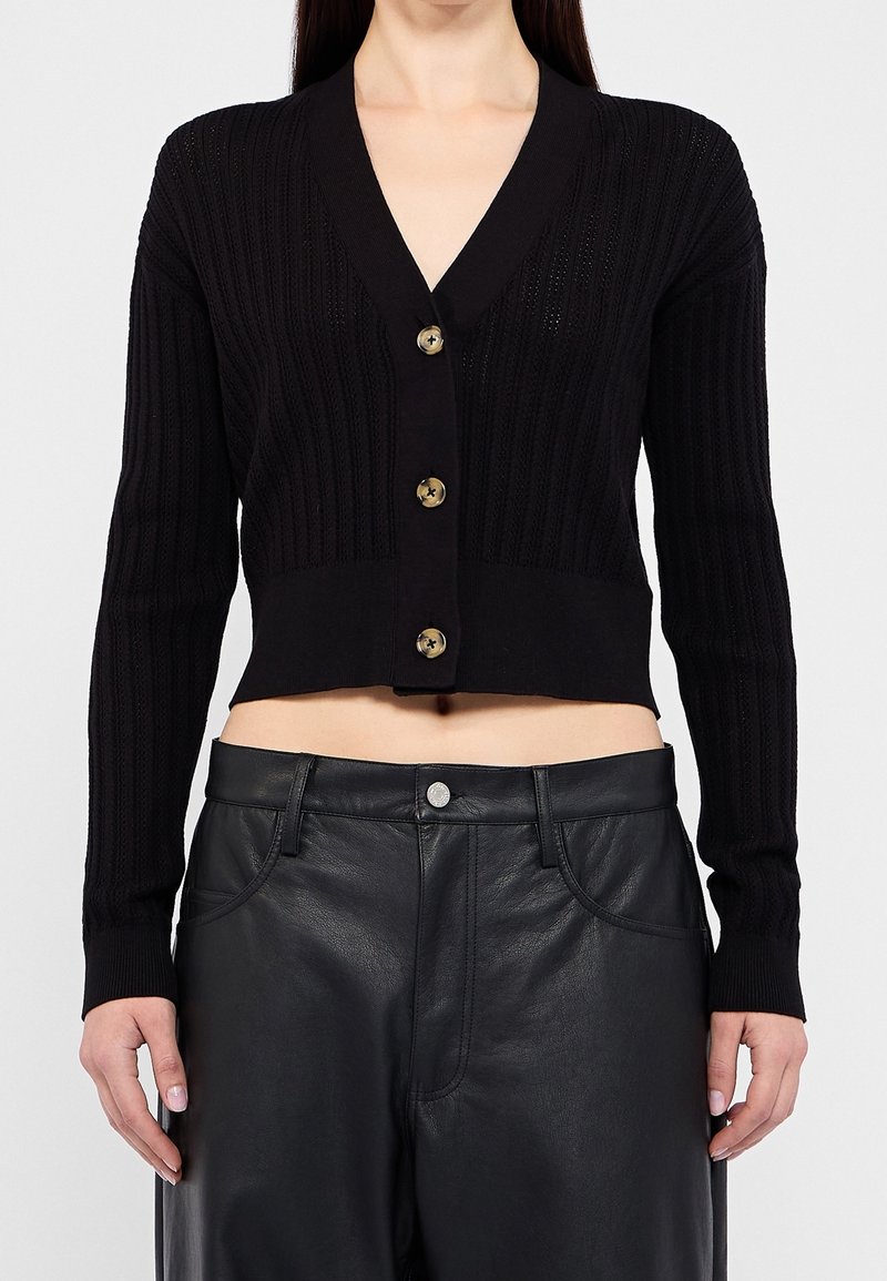Femme portant un cardigan court noir en côtes avec de gros boutons et un pantalon en cuir noir taille haute, debout devant un fond uni.
