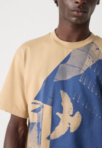 Beige t-skjorte med et stort blått grafikk som viser en fugl og geometriske mønstre. Myk stoff med rund hals og korte ermer.