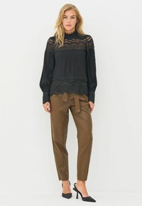 Czarna bluzka z koronką i wzorzystym dołem, z długimi rękawami i wysokim dekoltem, w połączeniu z brązowymi spodniami z sztucznej skóry i czarnymi szpilkami.