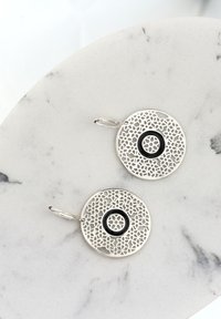 Silvercirkulära örhängen med intrikata spetsliknande utskärningar och en central svart accent. Hängande ögla för fäste. Texturerad yta.