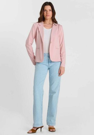 Mujer con blazer rosa claro, camiseta blanca, vaqueros azul claro y sandalias marrones de tacón, posando frente a un fondo blanco.