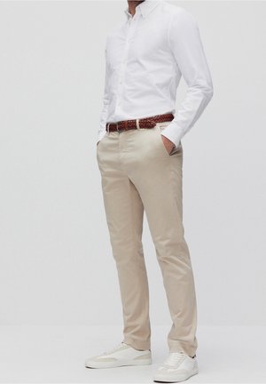 Homme portant un pantalon beige, une chemise blanche boutonnée, une ceinture tressée marron et des baskets blanches, debout les mains dans les poches.