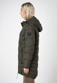 Frau im Profil, die eine lange, olivgrüne, gesteppte Jacke mit Kapuze und Reißverschlusstaschen vor einem schlichten, hellen Hintergrund trägt.