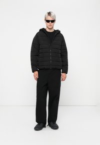 Doudoune noire avec des coutures horizontales, fermeture éclair à l'avant et capuche. Associée à un pantalon cargo noir et des chaussures noires avec des semelles texturées.