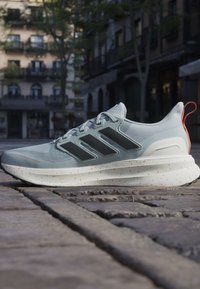 Lysegrå atletiksneaker med sorte striber og orange trækflig, placeret på brostensbelagt gade med slørede bygningskonstruktioner i baggrunden.
