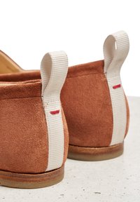 Brązowe zamszowe buty wsuwane z białymi tekstylnymi pętelkami, czerwonymi przeszyciami oraz teksturowaną podeszwą dla lepszej przyczepności.