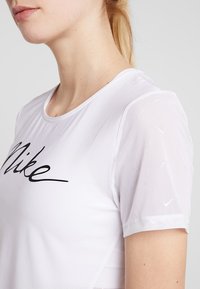 Camiseta atlética blanca con mangas cortas de malla, que presenta un logotipo negro de "Nike" en el pecho y sutiles patrones texturizados en las mangas.