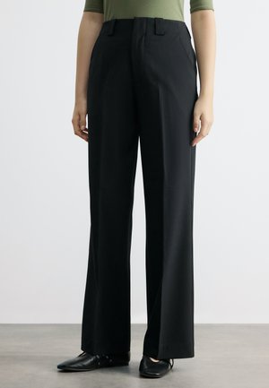 Άτομο που φοράει μαύρο παντελόνι φαρδιάς γραμμής, σετ με μαύρα μυτερά παπούτσια και ελιάς πράσινο τοπ, στέκεται σε γκρι πάτωμα μπροστά σε απλό φόντο.