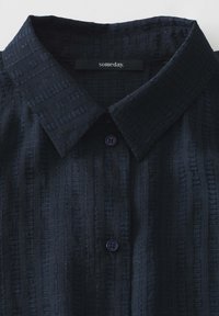 Camicia blu navy con un motivo tessuto, dotata di colletto e bottoni scuri. L'etichetta riporta "someday". Il tessuto ha una leggera lucentezza.
