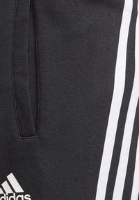 Pantalones de chándal negros con diseño de tres franjas blancas a lo largo de los lados, tejido texturizado y bolsillos laterales. Incluye el logo de Adidas en blanco.